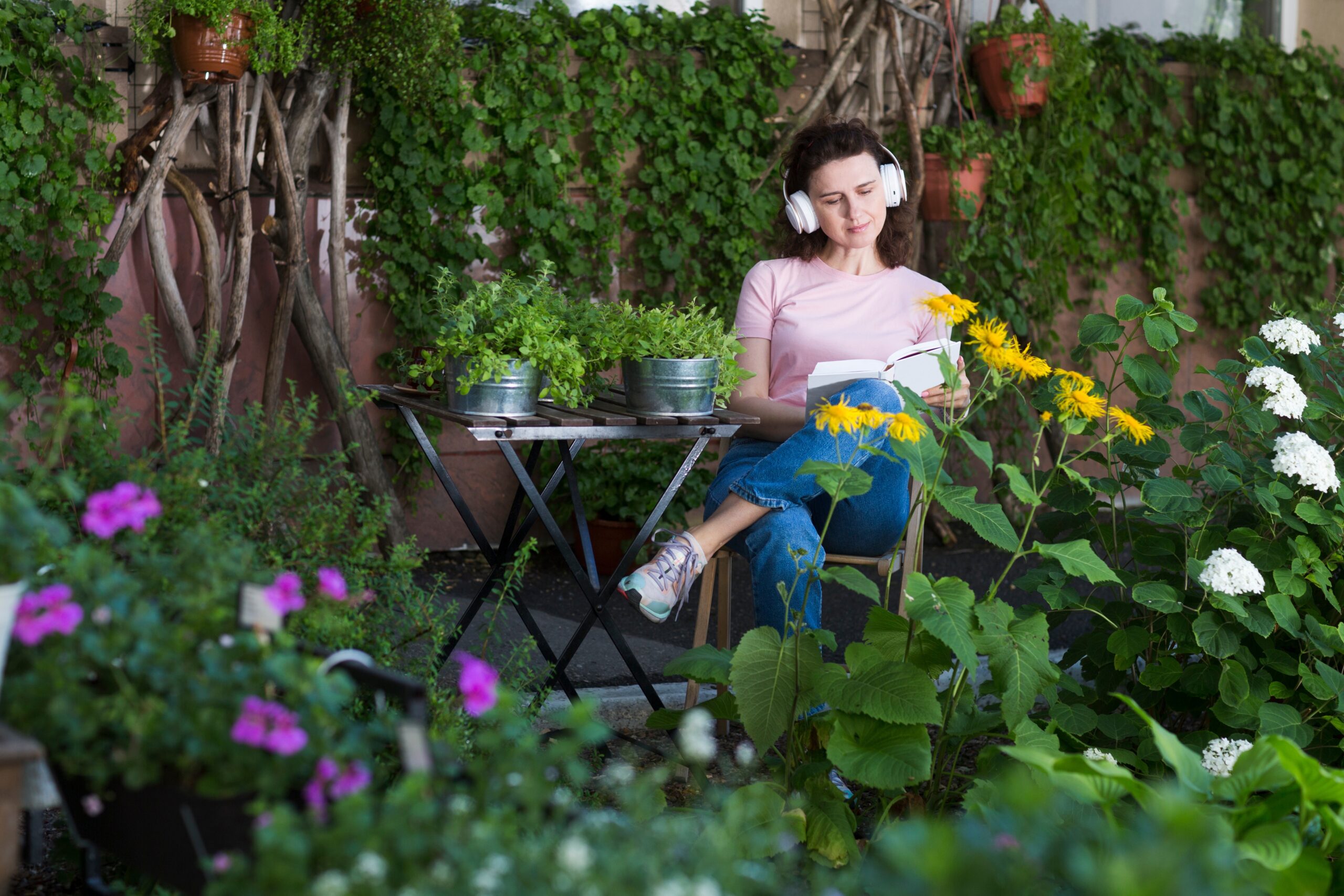 Kobieta w średnim wieku w słuchawkach czyta książkę i latem relaksuje się w ogrodzie niedaleko domu